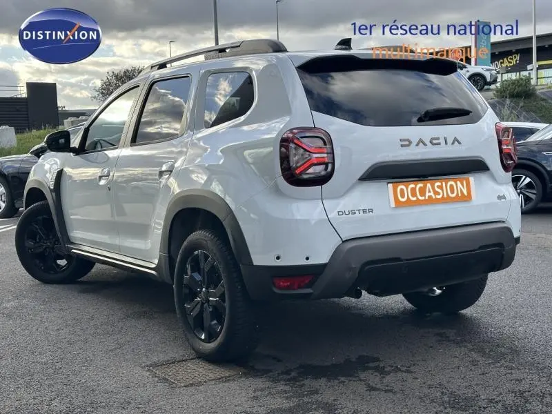 Vue 3/4 arrière droite du Dacia Duster blanc Glacier 2024 avec jantes noires et barres de toit visibles