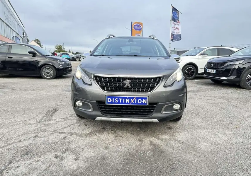 Vue frontale d'un Peugeot 2008 gris foncé Crossway avec calandre noire et feux allumés sur parking extérieur.