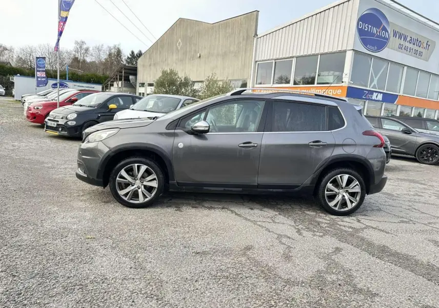 Profil latéral droit d'un Peugeot 2008 gris foncé avec jantes aluminium 17 pouces, stationné devant un bâtiment commercial.
