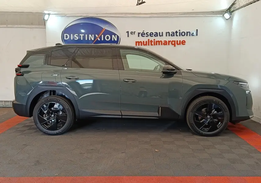 Vue de profil côté gauche d'une Citroën C5 hybride vert foncé avec jantes noires et toit noir en intérieur showroom.