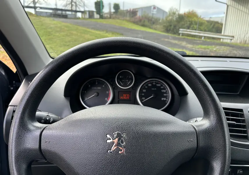 Vue intérieure centrée sur le volant noir de la Peugeot 206+ gris foncé, avec tableau de bord et compteurs visibles.
