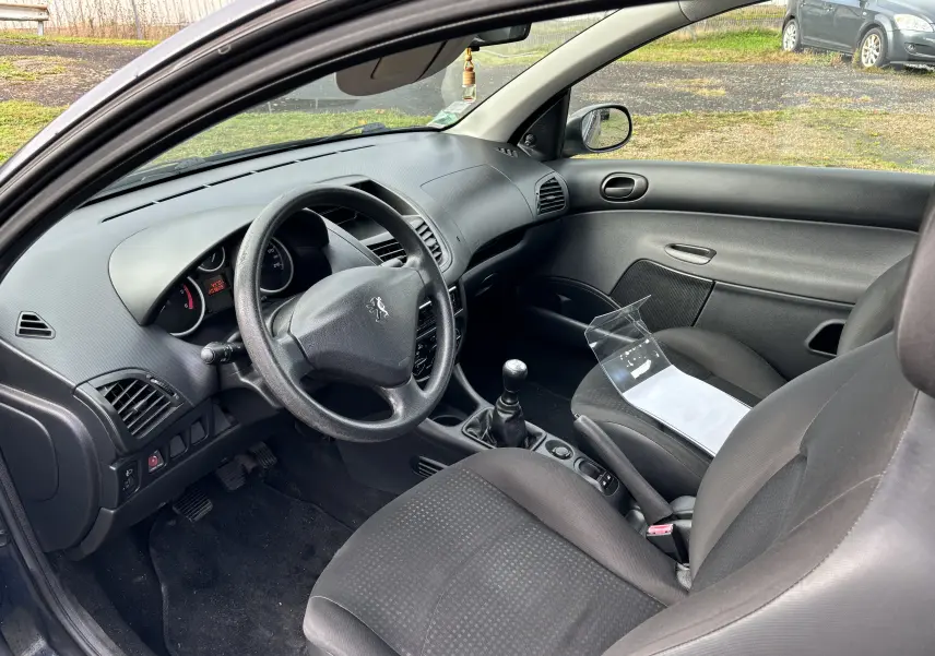 Intérieur côté conducteur d'une Peugeot 206+ gris foncé de 2009, volant, tableau de bord et levier de vitesses manuelle visibles.