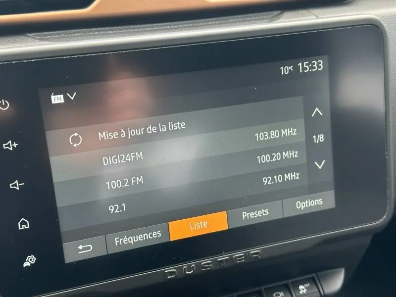 Écran tactile central du Dacia Duster 2024 blanc glacier affichant la liste des stations radio FM à 15h33, intérieur moderne.