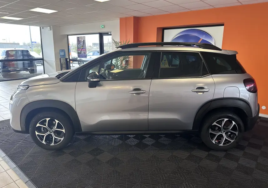 Vue de profil côté gauche d'une Citroën C3 Aircross 2024 gris Artense avec jantes alliage 16 pouces en intérieur showroom.