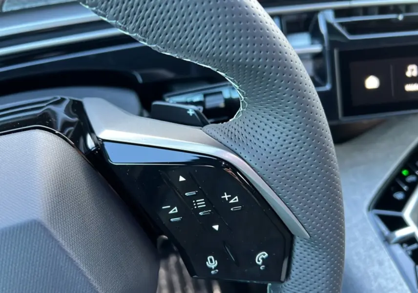 Gros plan sur le volant en cuir perforé gris avec commandes multifonctions, tableau de bord et écran tactile partiellement visibles.