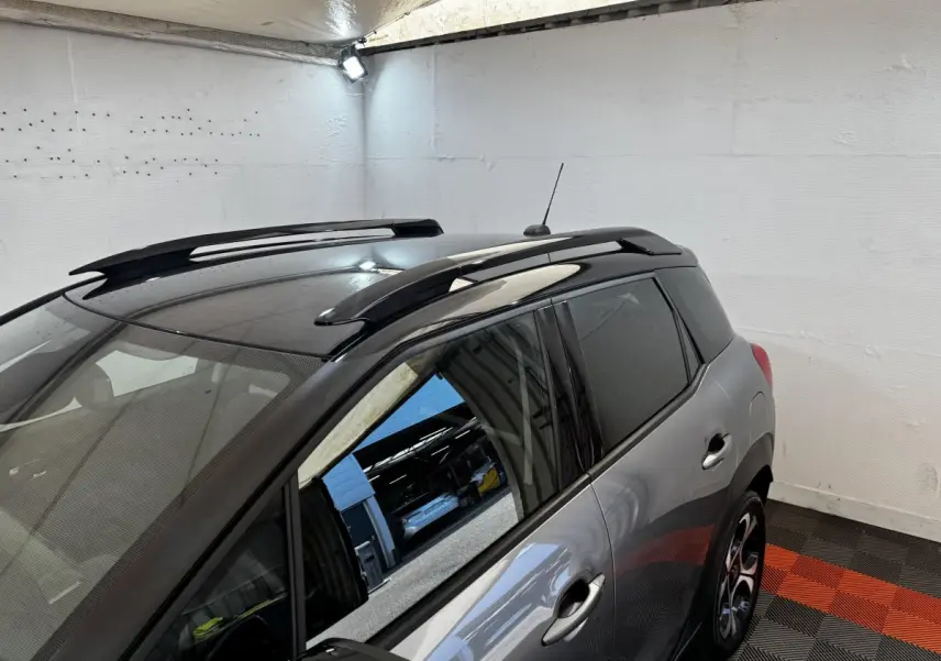Vue 3/4 arrière droite d'une Citroën C3 gris foncé avec toit noir et barres de toit en intérieur fermé.