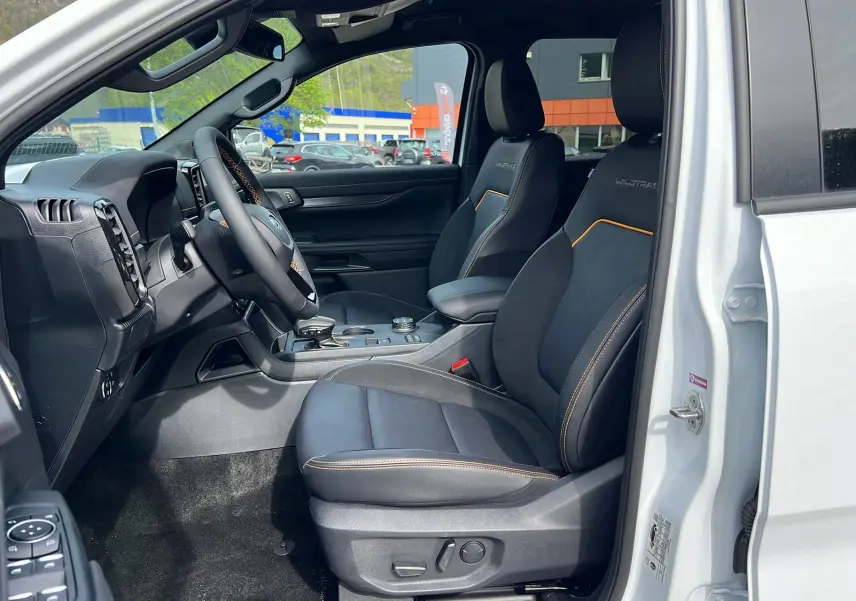 Vue intérieure côté conducteur du Ford Ranger blanc glacier 2025, sièges Wildtrak noirs avec surpiqûres orange et volant multifonctions.