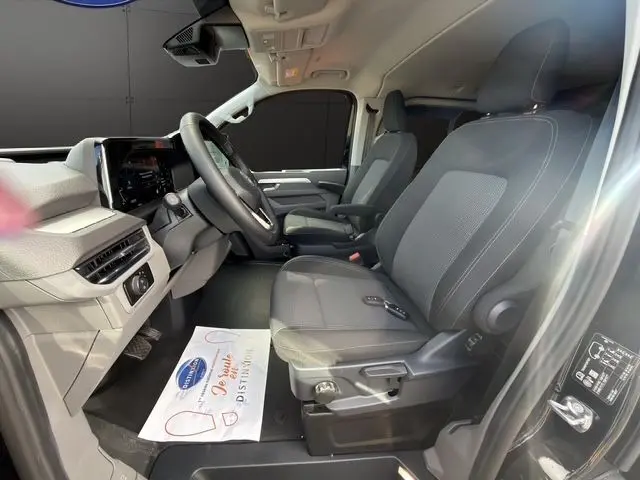 Vue intérieure côté conducteur du Volkswagen Transporter ProCab 2026, sièges avant en tissu rayé gris avec accoudoirs et volant multifonction.