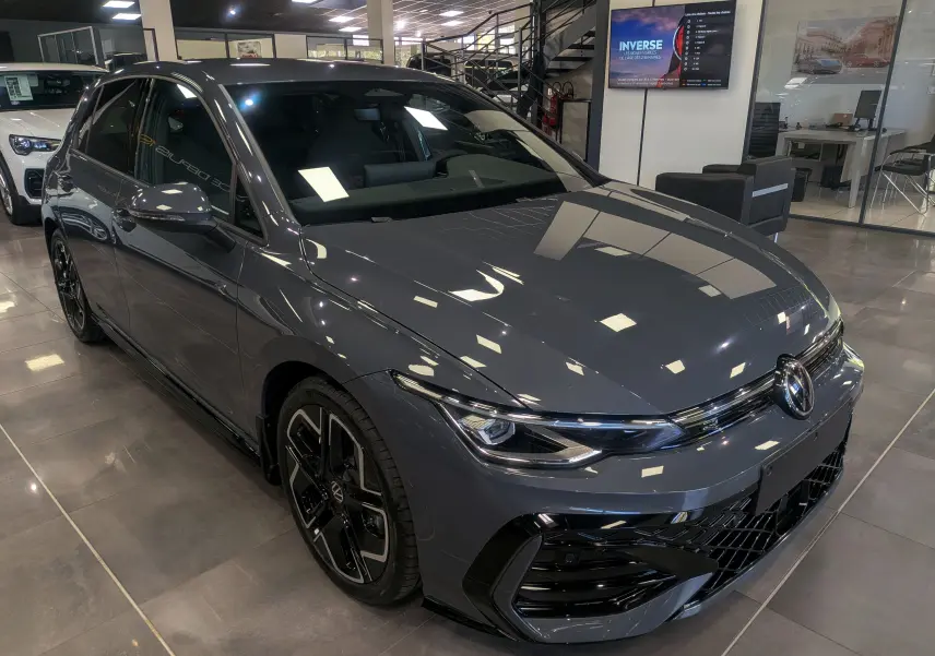 Vue 3/4 avant droite d'une Volkswagen Golf VIII 1.5 eTSI R-Line gris Urano avec jantes noires biton visibles en showroom.
