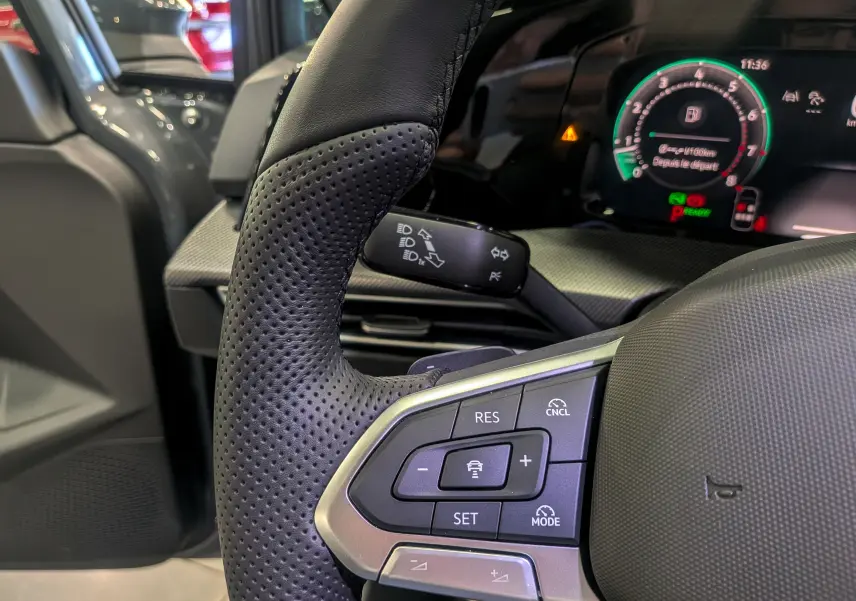 Gros plan sur le volant perforé côté gauche de la Volkswagen Golf VIII gris Urano avec commandes intégrées et tableau de bord numérique.