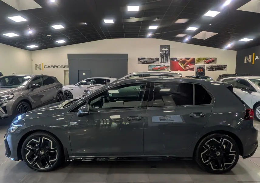 Vue de profil côté gauche d’une Volkswagen Golf VIII gris Urano, avec jantes noires et toit noir en showroom.