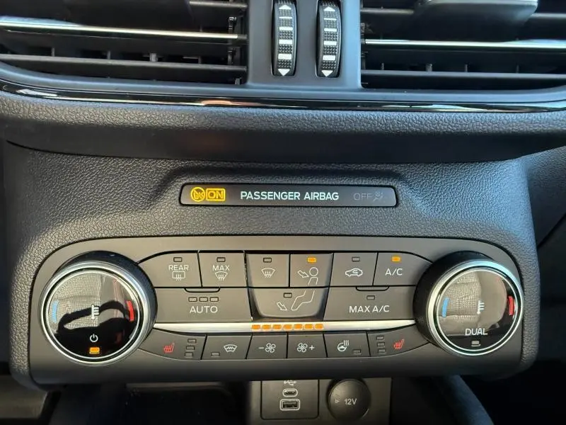 Vue rapprochée du tableau de bord du Ford Kuga 2024 noir, montrant les commandes de climatisation et l'indicateur airbag passager.
