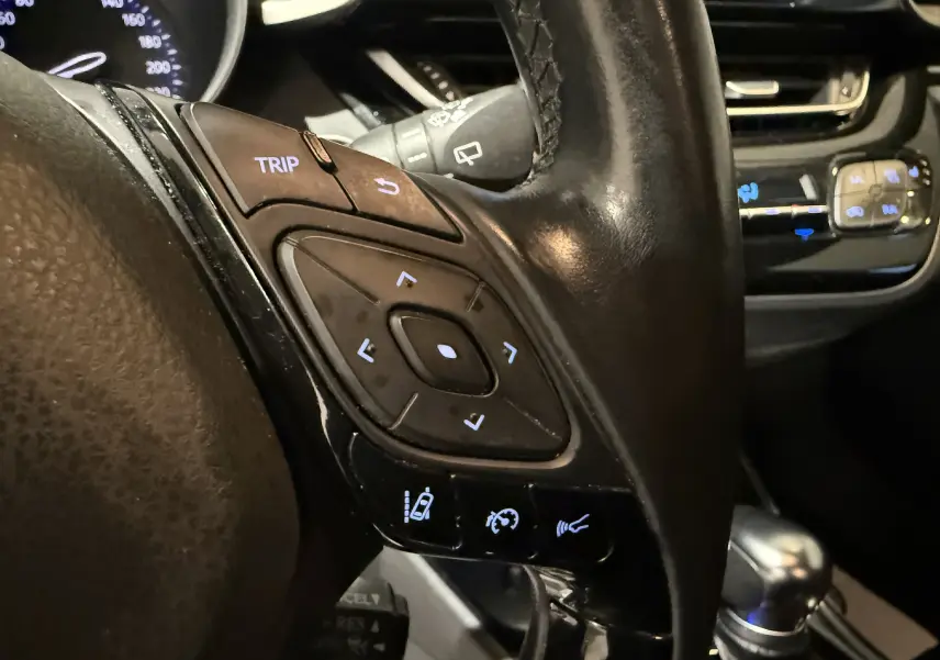 Gros plan sur les commandes du volant en cuir noir du Toyota C-HR Hybride rouge, avec boutons de régulateur et affichage lumineux.