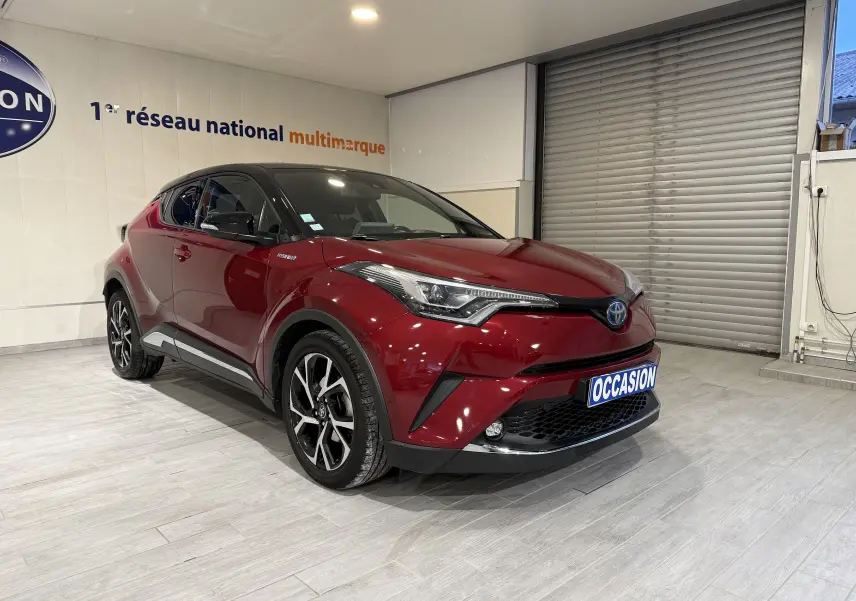 Vue 3/4 avant gauche d’un Toyota C-HR Hybride rouge brillant avec jantes noires et toit noir dans un showroom.
