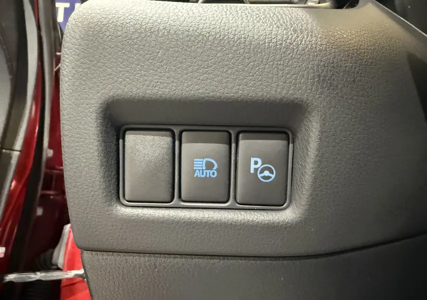 Gros plan sur les boutons de commande du tableau de bord d’un Toyota C-HR hybride rouge, avec éclairage automatique et assistance au stationnement.