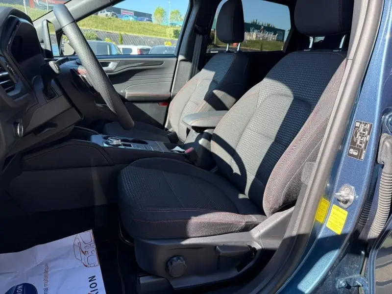 Vue intérieure côté conducteur du Ford Kuga bleu 2024, sièges sport en tissu noir avec surpiqûres rouges et volant cuir multifonction.