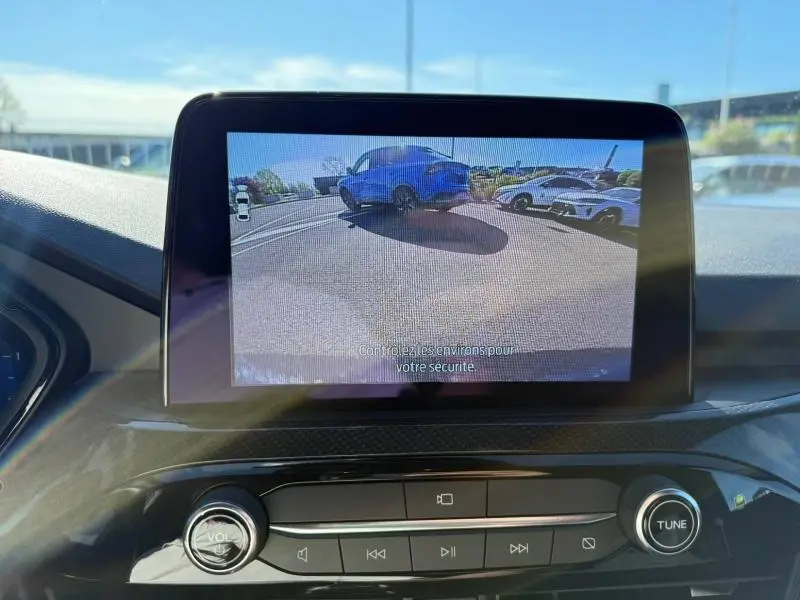 Écran central montrant la caméra de recul d’un Ford Kuga bleu 3/4 arrière dans un parking ensoleillé.