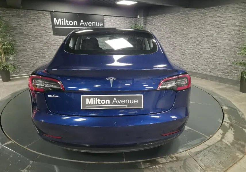 Vue arrière d'une Tesla Model 3 RWD bleu foncé avec feux LED et logo Tesla visible en intérieur showroom.