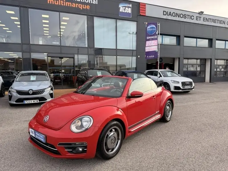 Volkswagen Coccinelle Cabriolet rouge vue 3/4 avant droit, capote beige relevée, devant un garage multimarque.