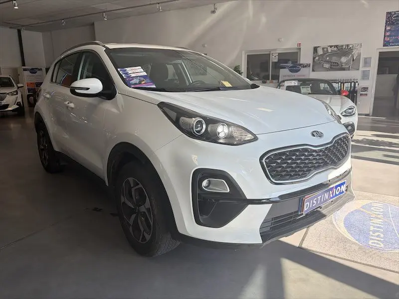Vue 3/4 avant gauche d'un KIA Sportage blanc 2021 en showroom, avec calandre noire et jantes bi-ton.