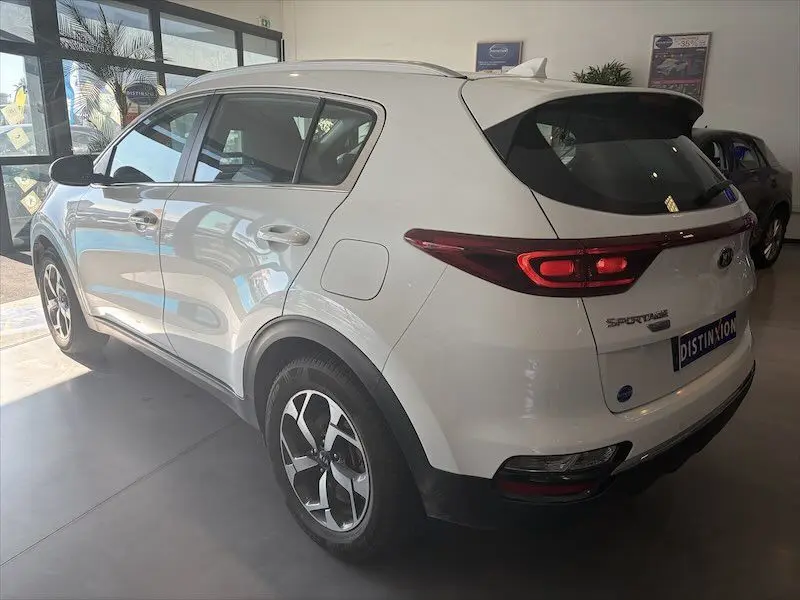 Vue 3/4 arrière droite d'un KIA Sportage blanc 2021 en intérieur, avec feux arrière allongés et jantes bi-ton.
