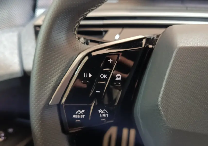 Gros plan sur les commandes multifonctions noires à gauche du volant perforé du Peugeot 5008 gris clair 2026.