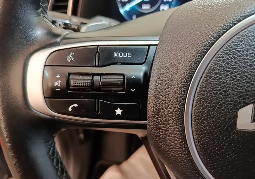 Gros plan sur les commandes gauche du volant cuir du Kia Sportage hybride rechargeable gris clair, avec affichage numérique en arrière-plan.