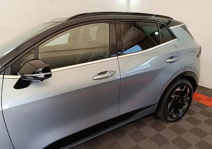Vue de profil côté gauche d’un KIA Sportage gris clair avec toit noir et jantes noires stylisées.