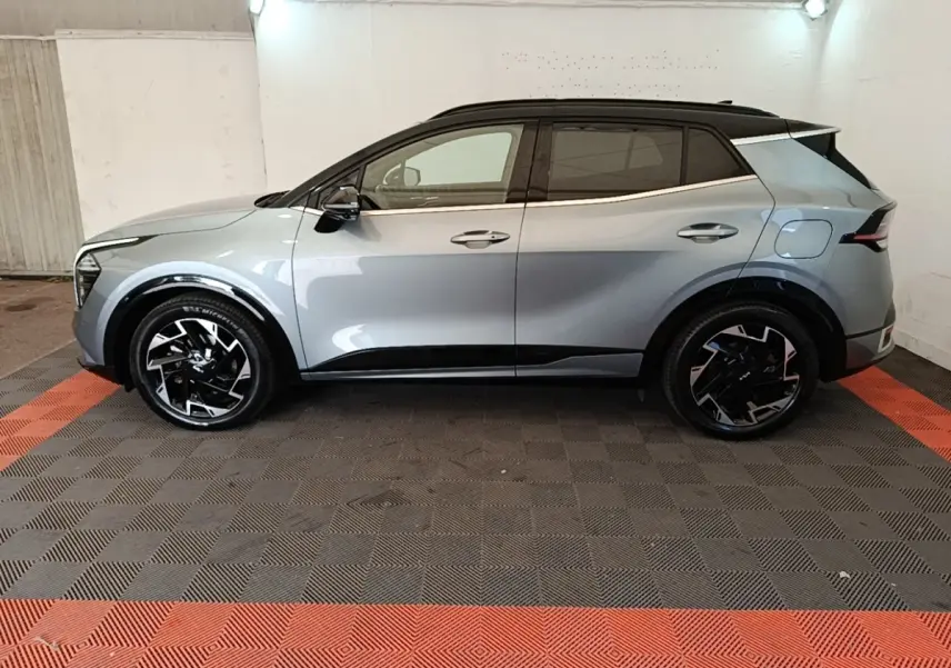 Vue de profil côté gauche d’un KIA Sportage gris clair avec toit noir et jantes noires à motifs argentés dans un garage.