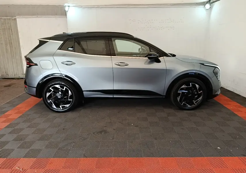 Vue de profil côté gauche d’un KIA Sportage gris clair avec toit noir et jantes noires stylisées dans un garage.