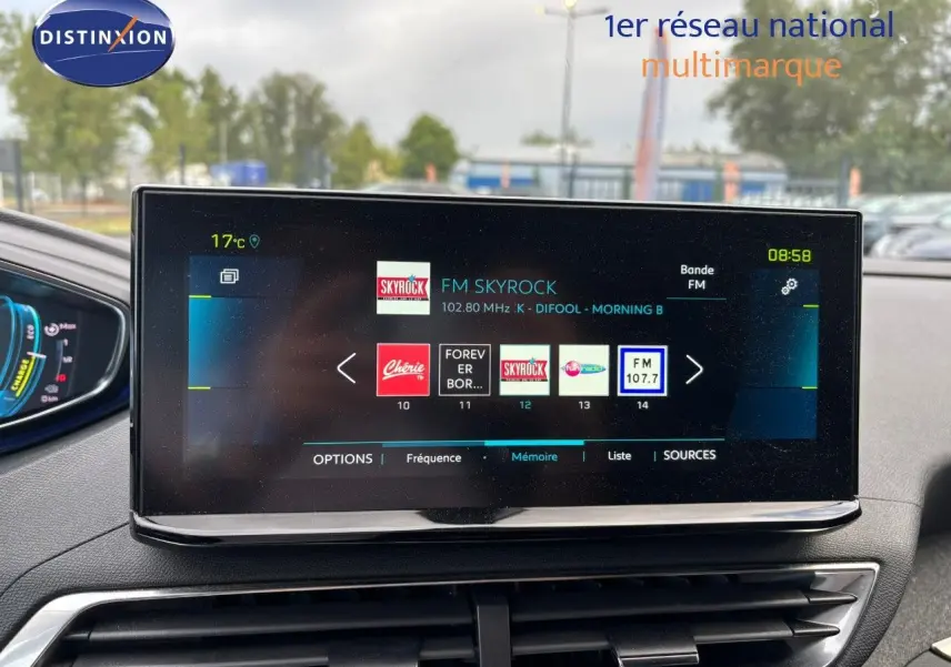 Écran tactile central du tableau de bord du Peugeot 3008 hybride, affichant les stations radio, avec vue sur l’extérieur flou.