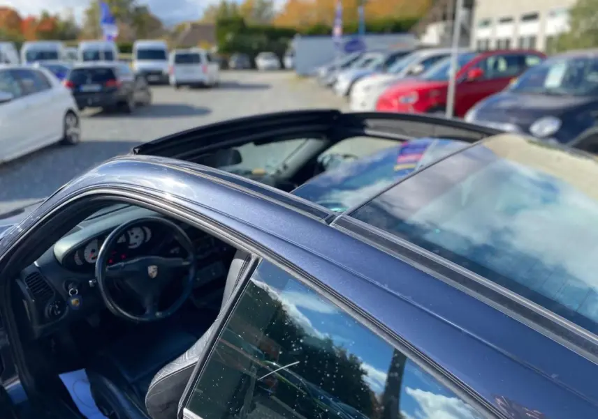 Vue côté gauche arrière d'une Porsche 911 Targa gris foncé avec toit coulissant ouvert et intérieur cuir noir visible.