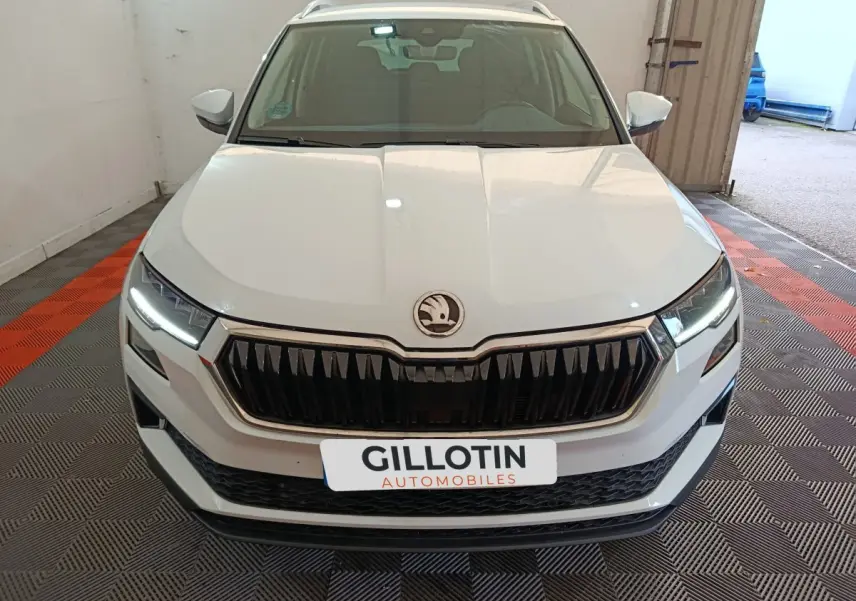 Vue avant d'un Skoda Karoq blanc 2024 avec calandre noire et phares LED allumés dans un garage.