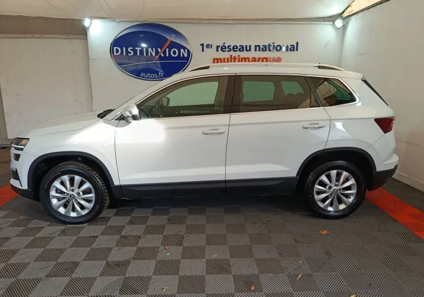 Vue latérale droite d'un Skoda Karoq blanc 2024 avec jantes alliage et barres de toit noires en intérieur showroom.