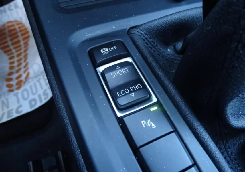 Gros plan sur les commandes de modes de conduite et assistance au freinage dans l’habitacle noir du BMW X1 blanc.