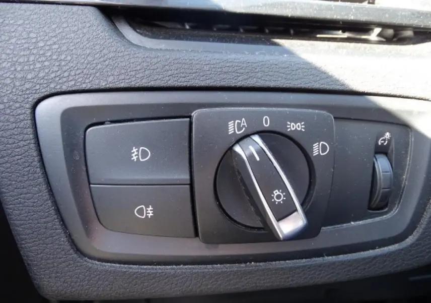 Gros plan sur le sélecteur de feux noir du tableau de bord du BMW X1 blanc, avec commandes d'éclairage et réglage de luminosité.