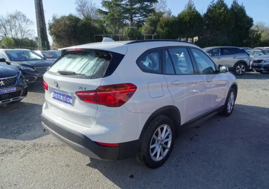 BMW X1 blanc vue 3/4 arrière droit sur parking, avec hayon et antenne aileron de requin visibles.