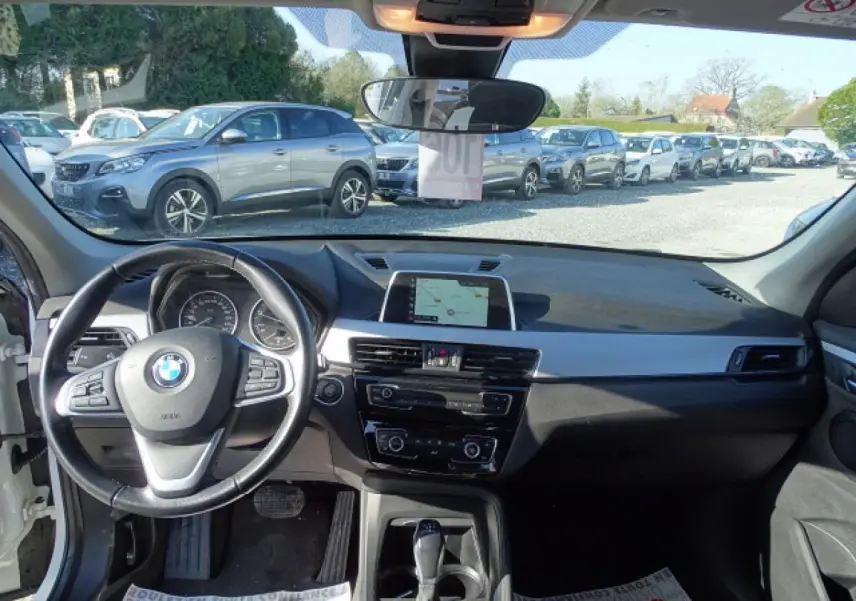 Vue intérieure avant de la BMW X1 blanche 2017, montrant le volant cuir multifonction et l’écran de navigation central.