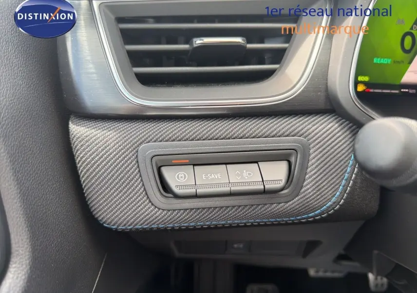 Gros plan sur les commandes E-SAVE et réglage des phares, tableau de bord avec finition carbone et surpiqûres bleues du Renault Symbioz gris.