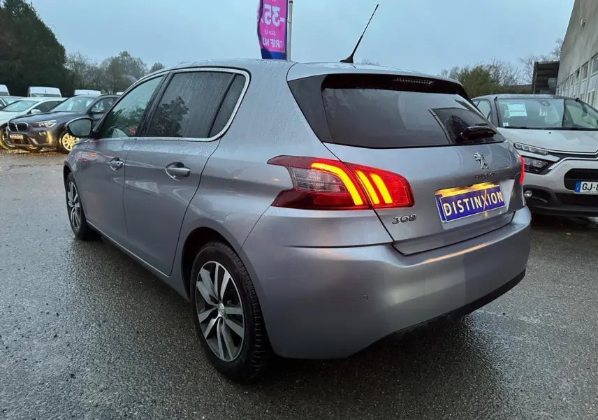 Vue 3/4 arrière gauche d'une Peugeot 308 gris Artense avec feux arrière allumés sur parking humide.