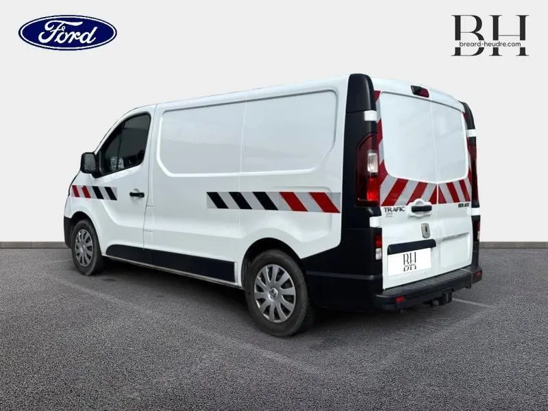 Vue 3/4 arrière droite d’un Renault Trafic Fourgon blanc Glacier avec bandes réfléchissantes rouges et noires et attelage de remorque.