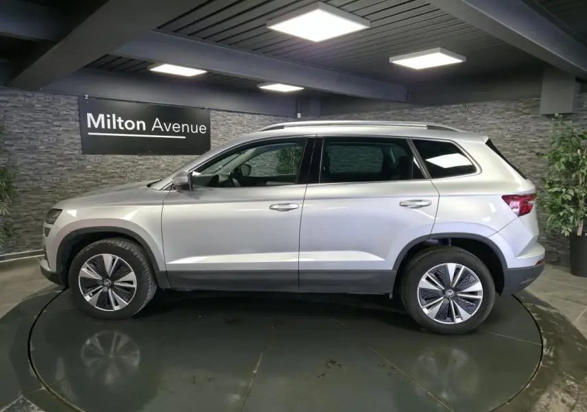 Vue latérale droite d'un SUV Skoda Karoq gris 2023 avec jantes alliage et barres de toit argentées dans un showroom.
