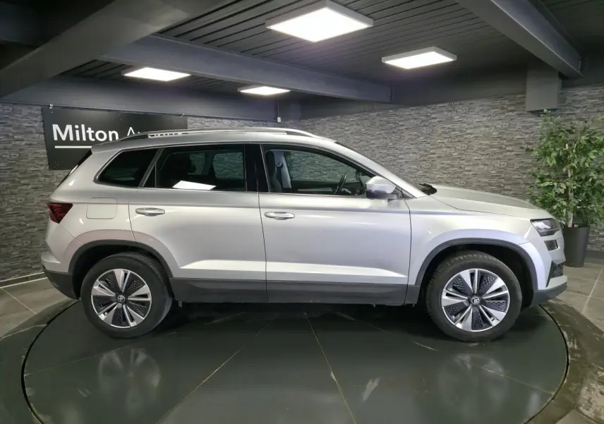 Vue latérale droite d'un SKODA KAROQ gris 2023 en intérieur avec jantes alliage et barres de toit noires.