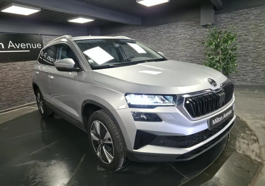 Skoda Karoq gris en 3/4 avant droit, phares allumés et calandre noire brillante dans un showroom.