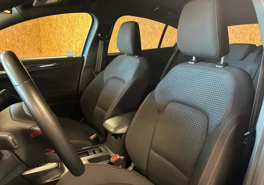 Vue intérieure côté avant gauche de la Ford Focus 2024, sièges tissu noir avec surpiqûres rouges et volant cuir perforé.