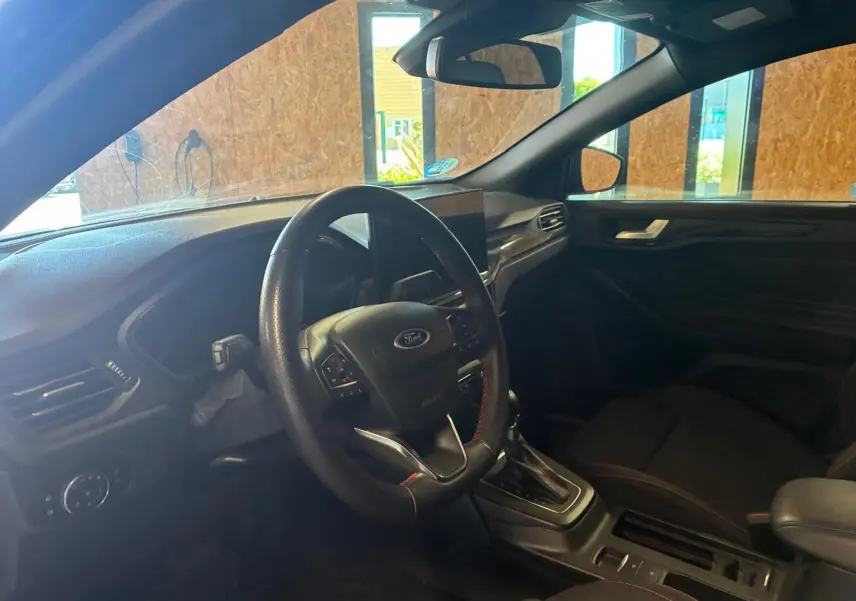 Vue intérieure côté conducteur de la Ford Focus 2024, tableau de bord et volant noir avec surpiqûres rouges.