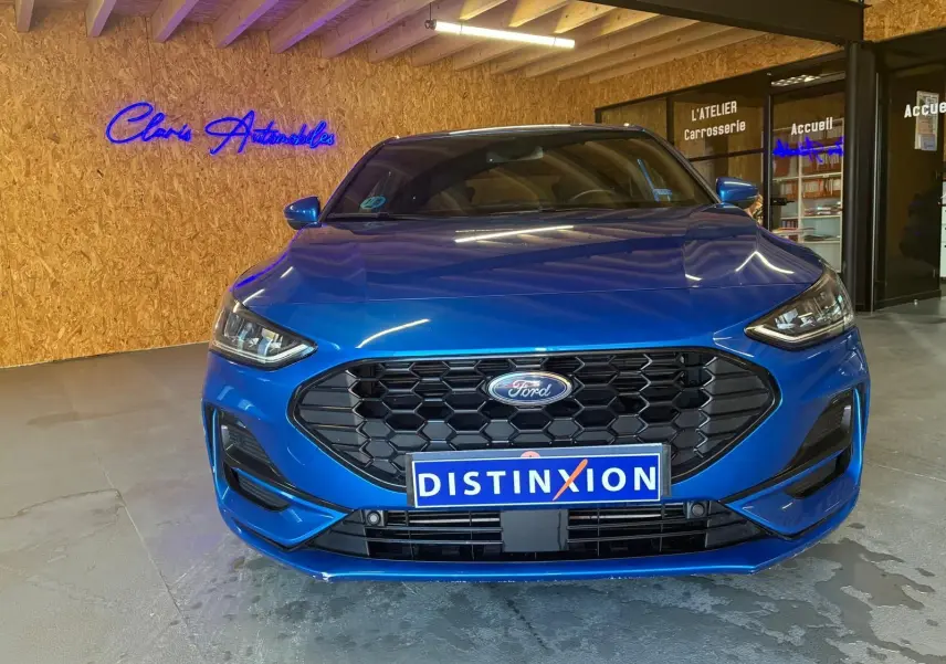 Vue frontale d'une Ford Focus bleue 2024 avec calandre noire hexagonale et plaque Distinxion en intérieur d'atelier.