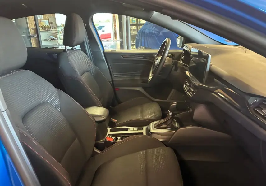 Vue intérieure côté gauche de la Ford Focus 2024 bleue, montrant sièges tissu noirs avec surpiqûres rouges et tableau de bord moderne.