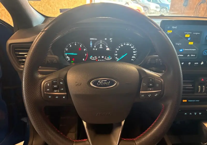 Vue rapprochée du volant noir avec surpiqûres rouges et tableau de bord numérique de la Ford Focus 2024 bleu hybride.