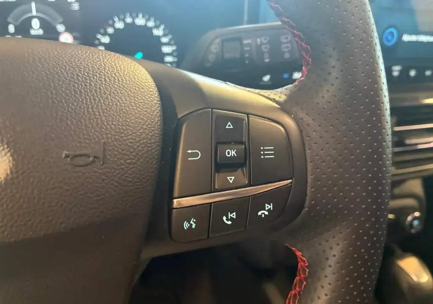 Gros plan sur le volant perforé avec surpiqûres rouges de la Ford Focus 2024, boutons de commande multifonctions visibles.